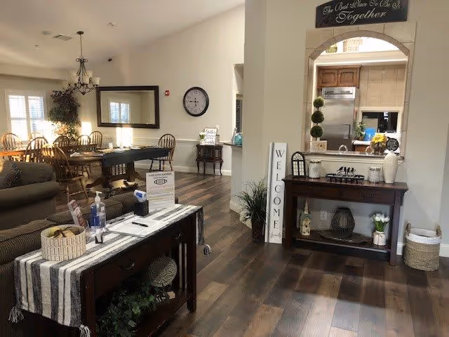 Interior view of a senior living facility showing a spacious common area with wooden flooring. There is a dining area with wooden chairs and tables on the left side, a comfortable seating area with sofas, and a console table with hand sanitizer and informational materials in the foreground. A decorative table with plants and a 'WELCOME' sign is positioned near an arched opening leading to a kitchen area. The walls are light-colored, and the space is well-lit with natural light coming through windows with white blinds.