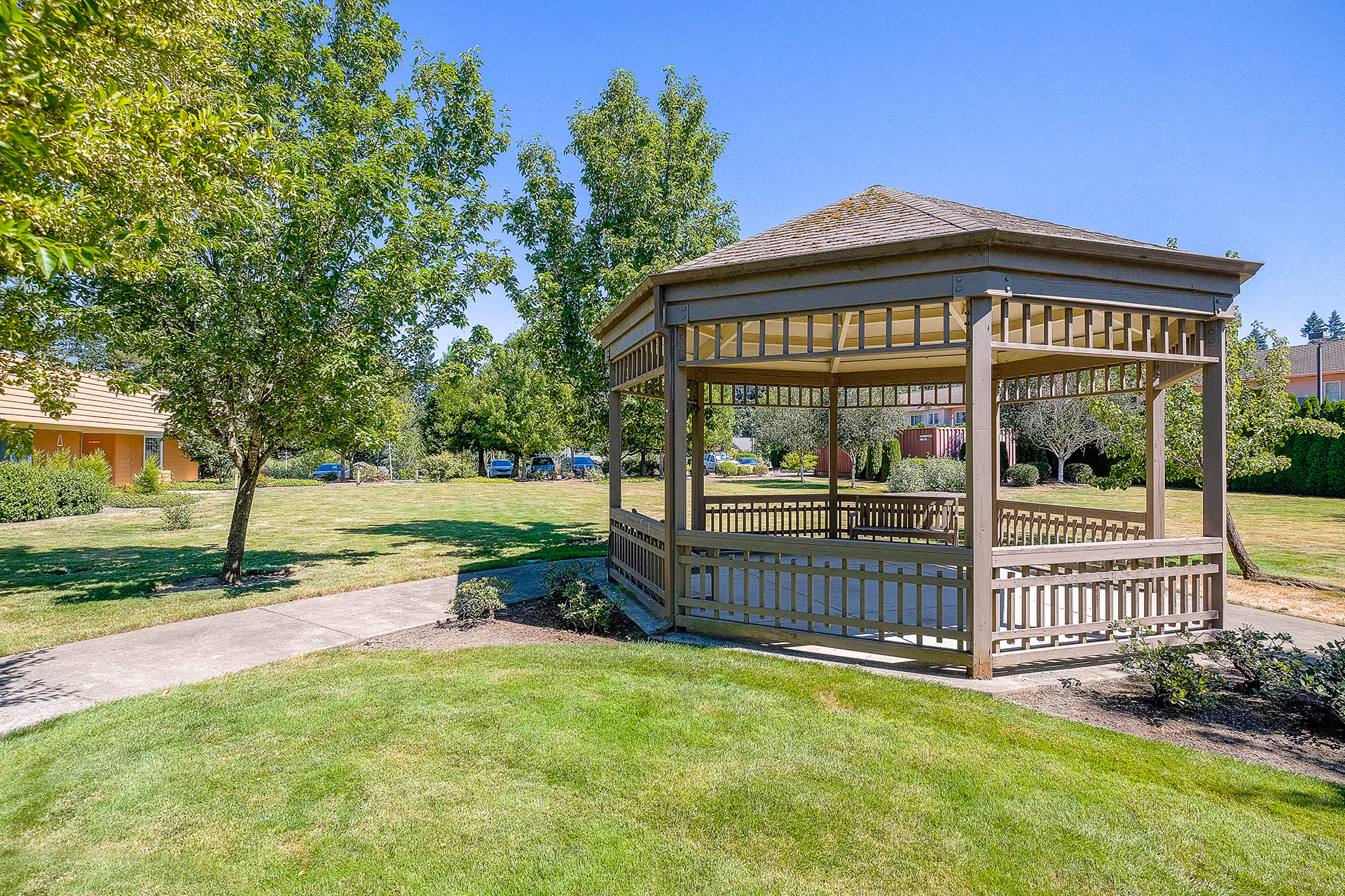 A wooden gazebo situated in a well-maintained grassy outdoor area with trees and shrubs under a clear blue sky. A paved walkway curves around the gazebo, and buildings are visible in the background.