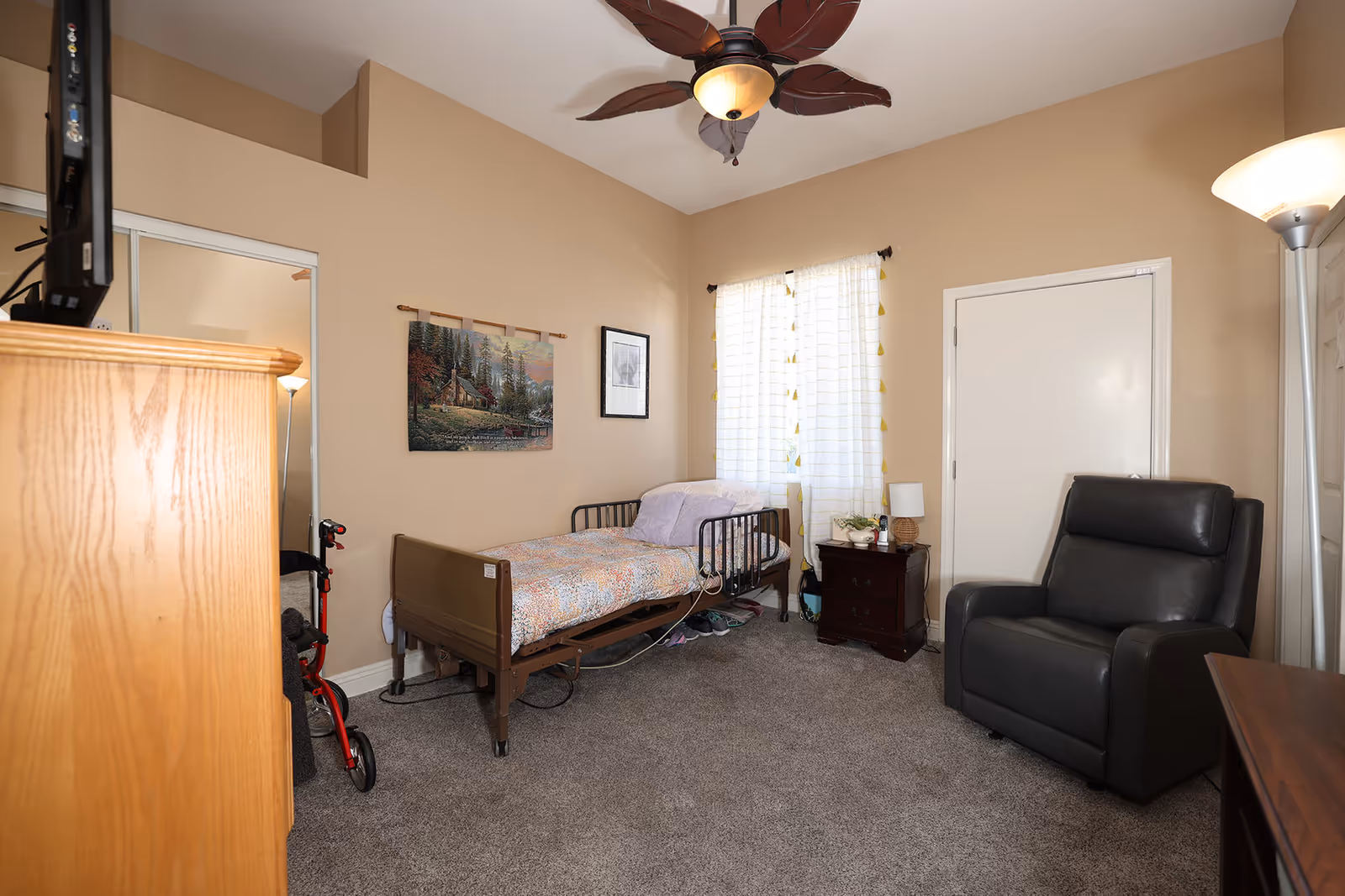 A senior living bedroom with a hospital-style bed against a beige wall, a wooden dresser with a TV on top, a black recliner chair, a nightstand with a lamp and some items, a window with white curtains, a ceiling fan with leaf-shaped blades, and a walker partially visible near the dresser.