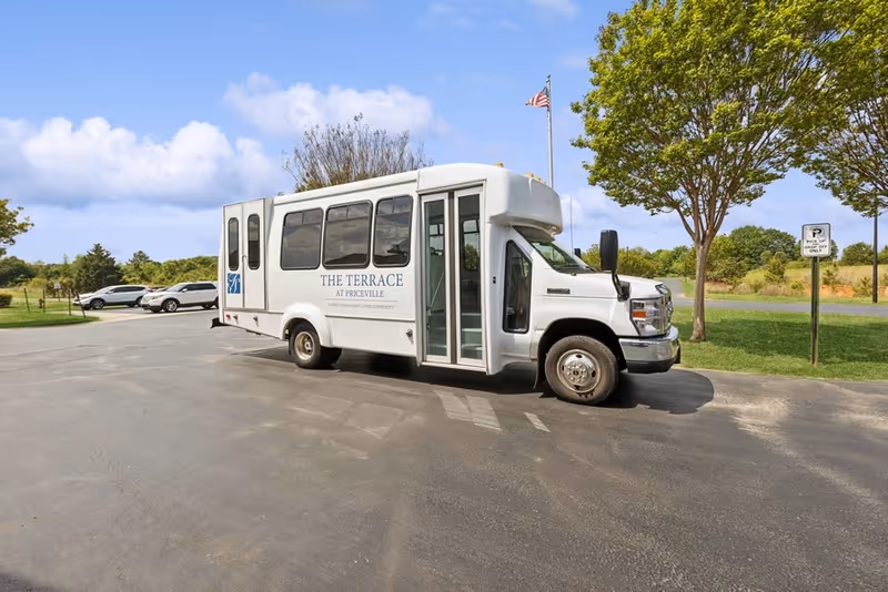 A white shuttle bus labeled "The Terrace at Priceville" parked in a lot by trees and an American flagpole.