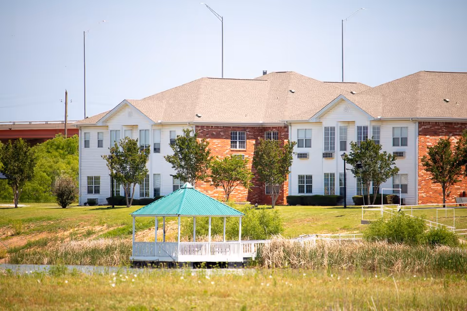 Two-story brick-and-white assisted living building with trees and a green-roofed gazebo by a small pond on a grassy lawn.