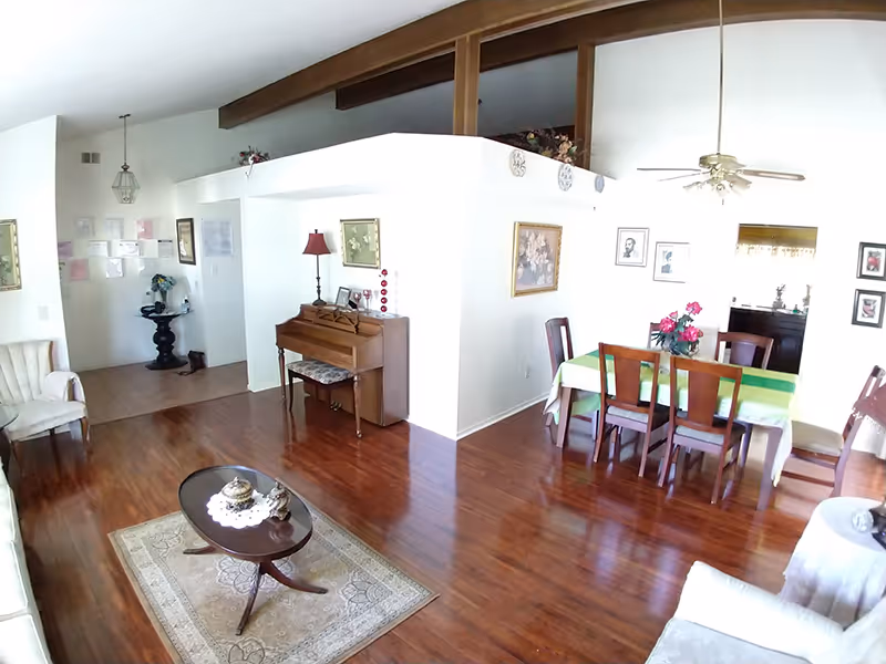 Open living and dining area with hardwood floors, a small piano, coffee table and rug, and a dining table under a vaulted ceiling with exposed beams.