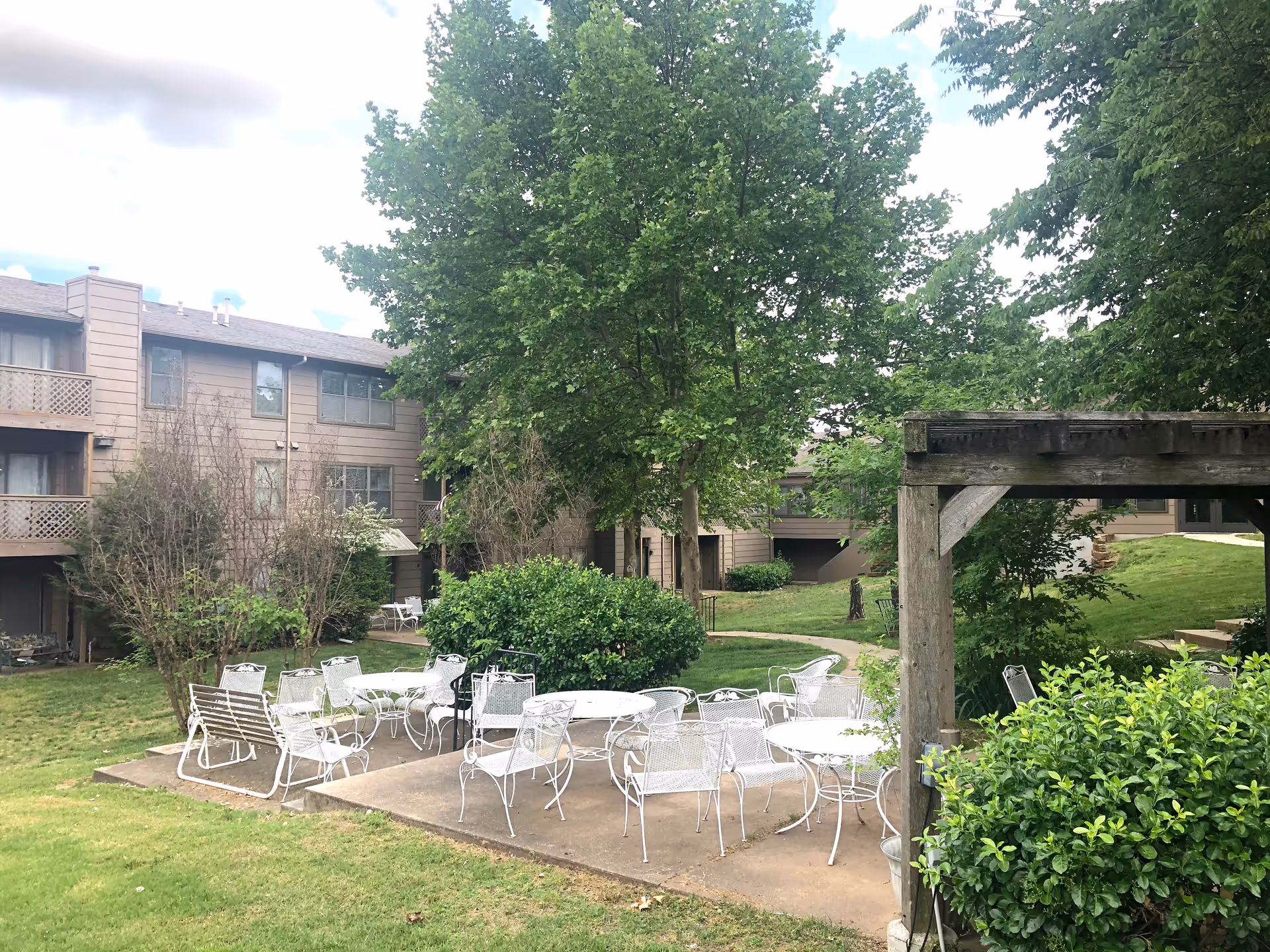 Outdoor patio area with multiple white metal tables and chairs arranged on a concrete slab surrounded by green grass, bushes, and trees. In the background, there is a multi-story residential building with balconies and windows.