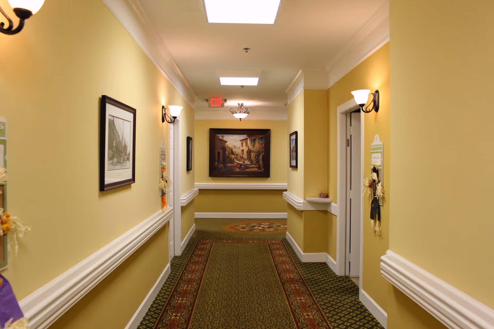 A well-lit hallway in a senior living facility with yellow walls, green patterned carpet, framed artwork on the walls, and wall-mounted light fixtures. Doors line the hallway, and there is an exit sign visible at the end.
