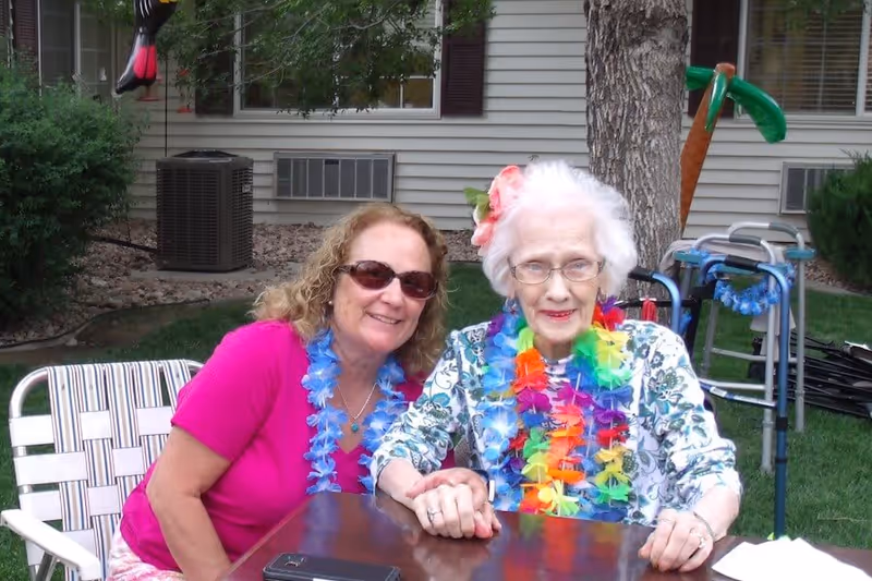 Two women sitting outdoors at a table in a garden area. One woman is elderly with white hair, wearing glasses, a floral top, and colorful leis. The other woman has curly hair, sunglasses, and a pink shirt, also wearing a lei. There is a walker and some greenery in the background.
