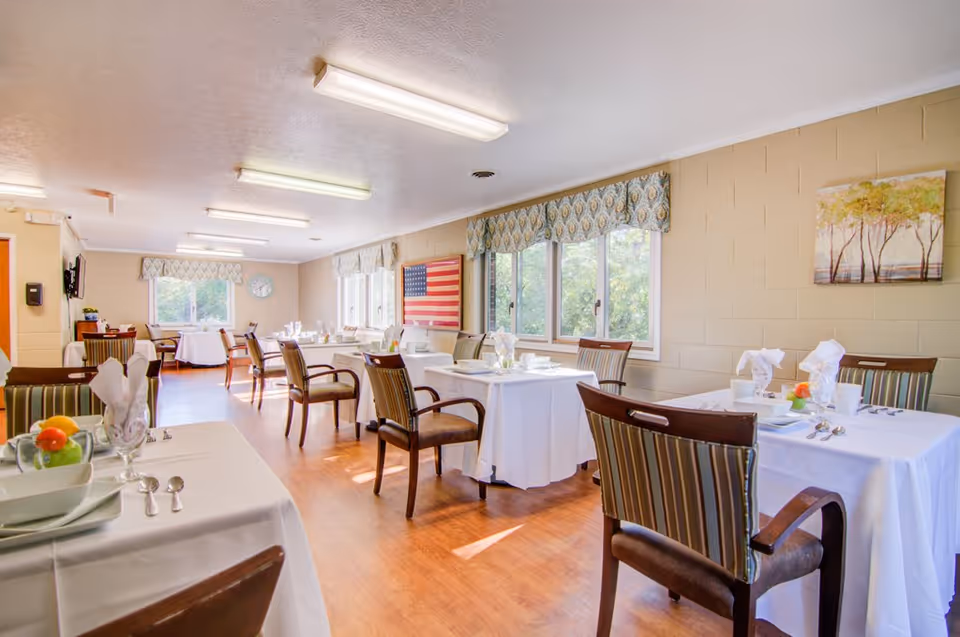 A bright dining room in a senior living facility with several tables covered in white tablecloths, each set with plates, bowls, utensils, and napkins folded in a decorative manner. The room has large windows with patterned valances letting in natural light, wooden chairs with striped upholstery, and a wooden floor. On the wall, there is an American flag and a painting of trees.
