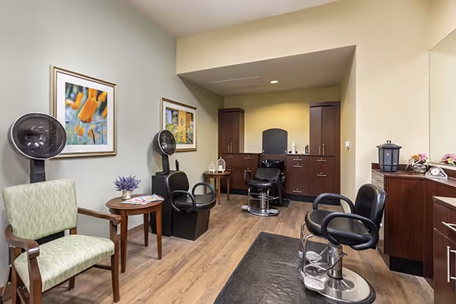 Interior view of a hair salon area in a senior living facility with salon chairs, hair dryers, wooden cabinets, a small table with magazines, and framed artwork on the walls.