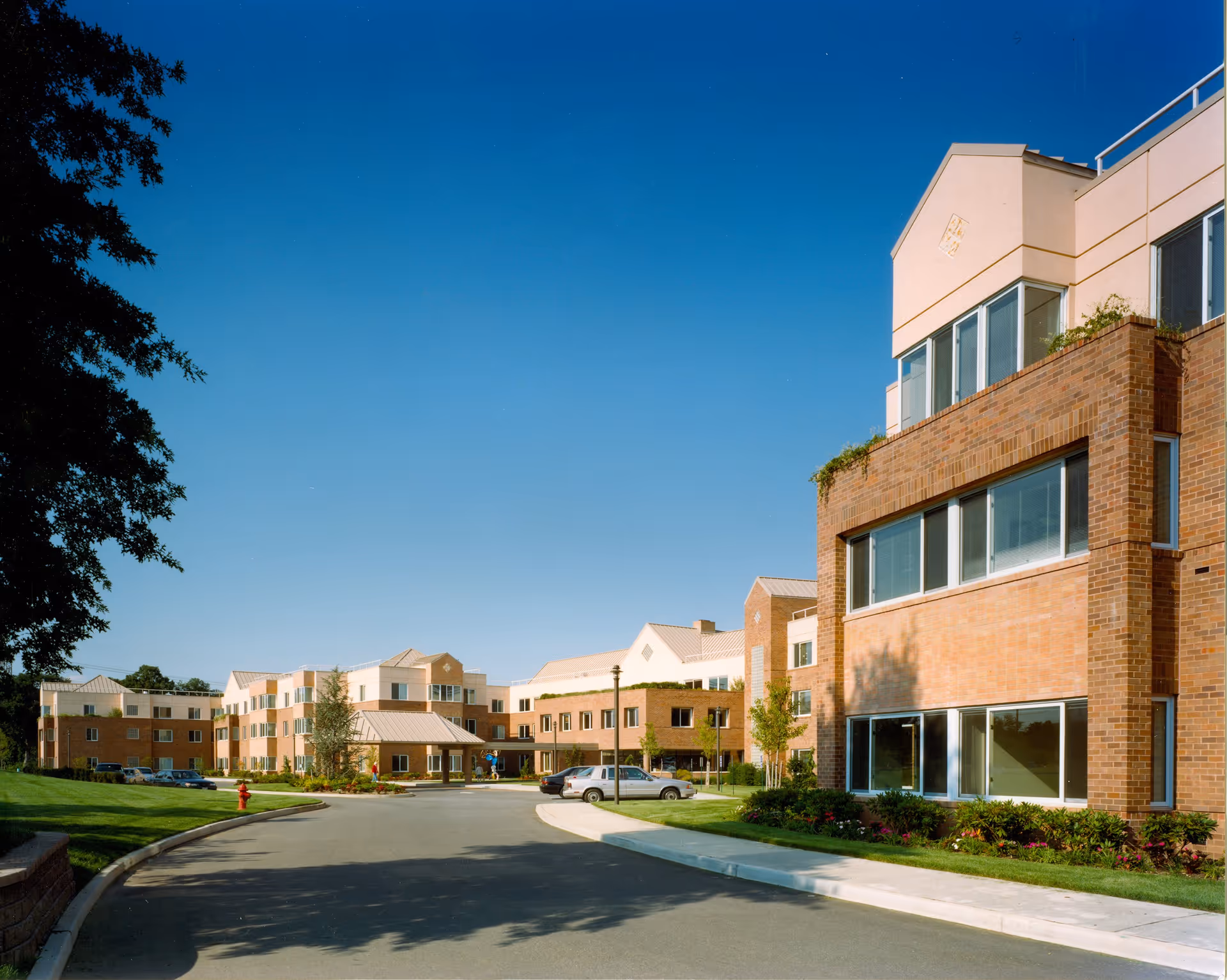 Exterior view of a multi-story senior living facility building with brick and light-colored walls under a clear blue sky. There is a driveway with parked cars and landscaped green areas with trees and shrubs around the building.