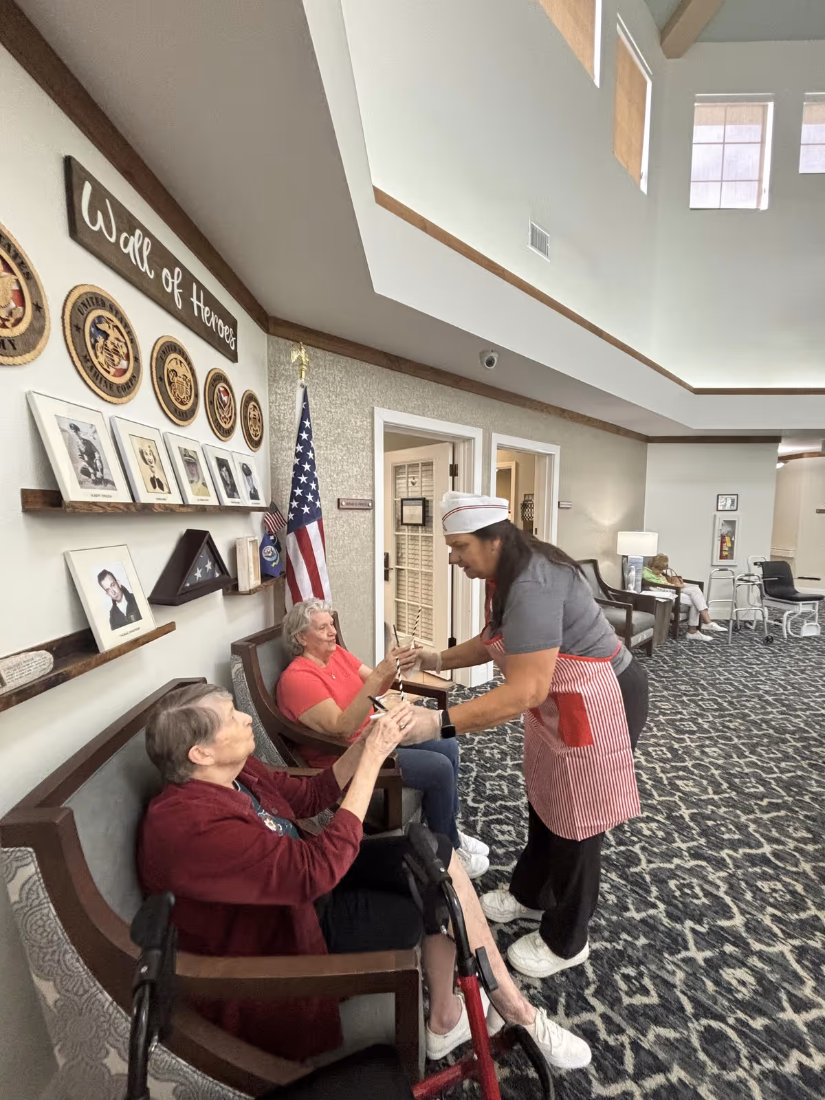 A caregiver wearing a red and white striped apron and hat is handing a small item to an elderly woman seated in a chair with a walker in front of her. Another elderly woman in a red shirt is seated next to her. The setting is a spacious, well-lit common area with a 'Wall of Heroes' display featuring military emblems and framed photos, and an American flag in the background. Additional seating and a person sitting further back are visible.
