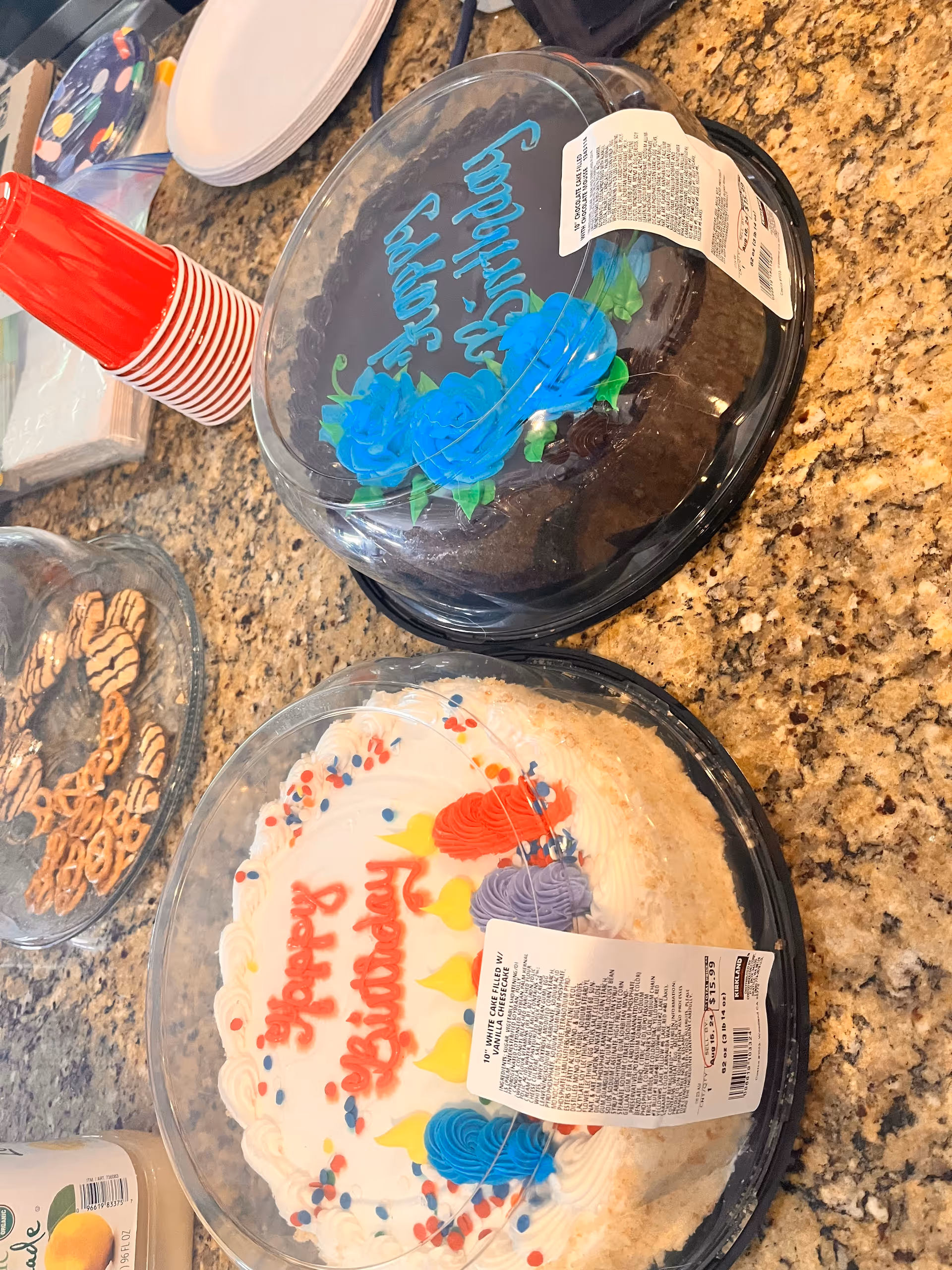 Two birthday cakes in plastic containers on a granite countertop. One cake has white frosting with colorful decorations and the words 'Happy Birthday' written in red. The other cake has chocolate frosting with blue flower decorations and the words 'Happy Birthday' written in blue. Nearby are red plastic cups, a plate of pretzels, and some paper plates and napkins.