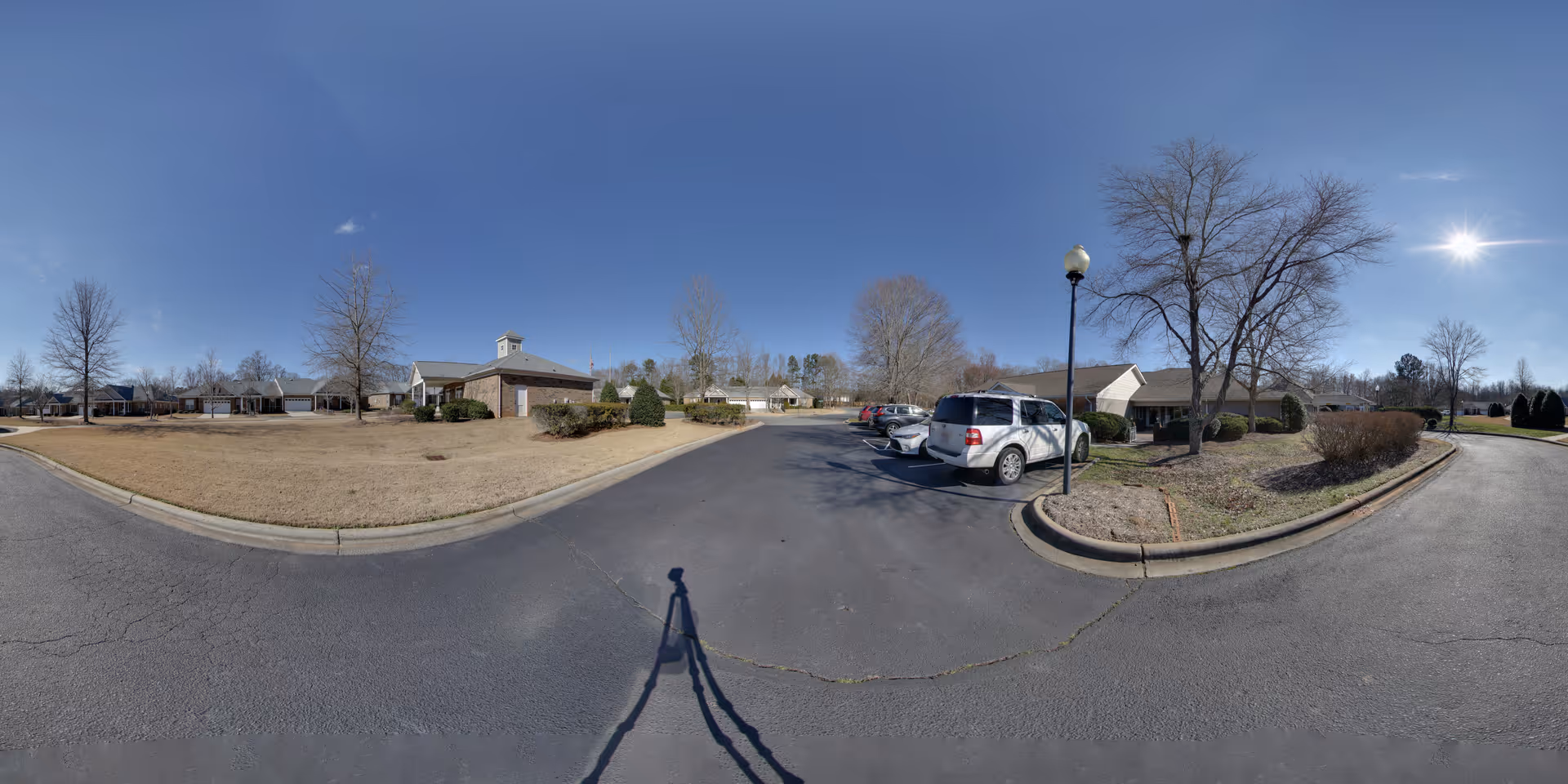 Panoramic exterior view of a senior living community showing low brick buildings, a parking lot with cars, landscaped lawns, and bare trees under a clear blue sky.