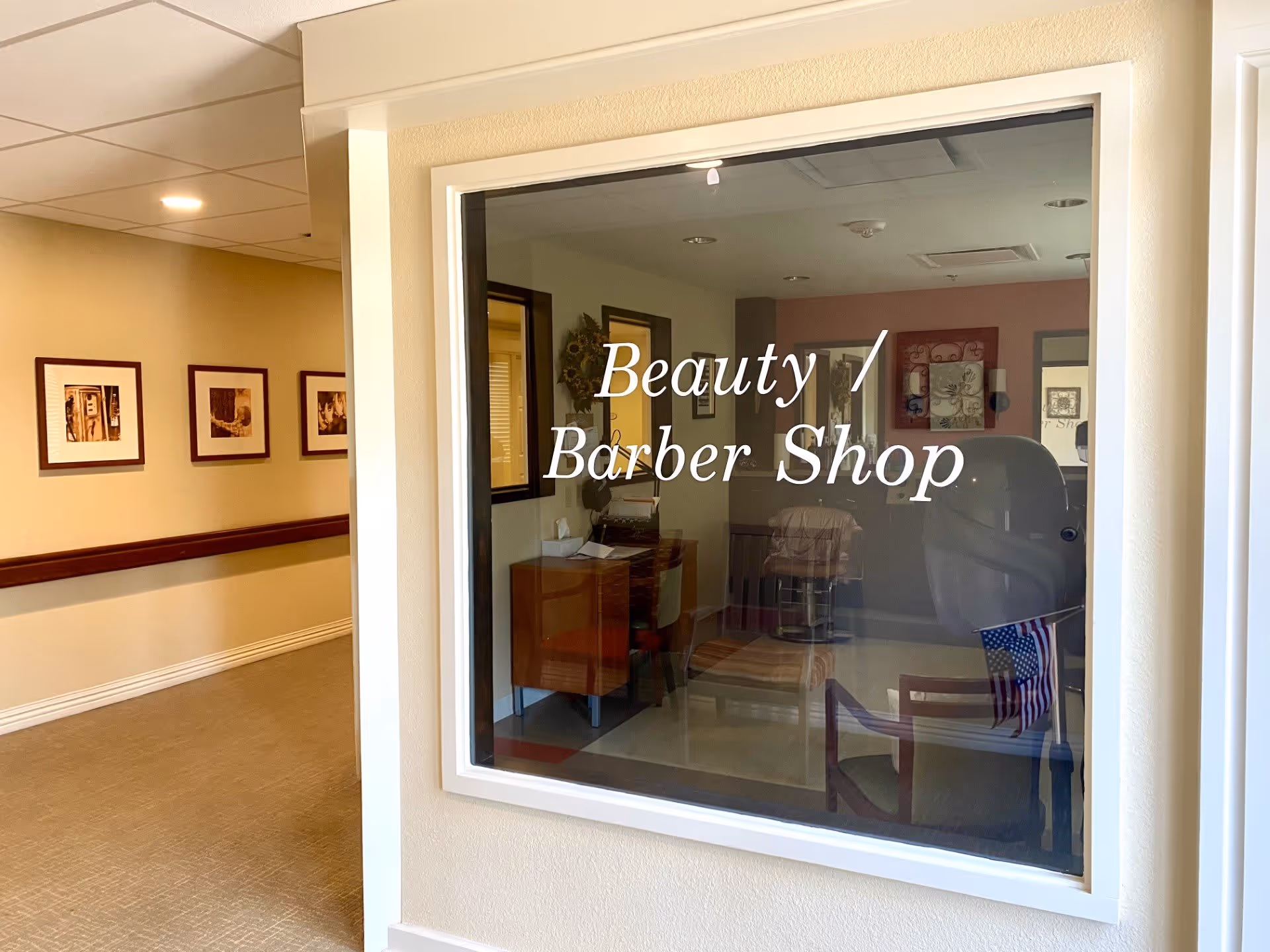 Interior view of a hallway in a senior living facility with a large window showing a beauty and barber shop. The shop contains chairs and a small desk, and the window has the text 'Beauty / Barber Shop' written on it. The hallway walls are decorated with framed pictures.