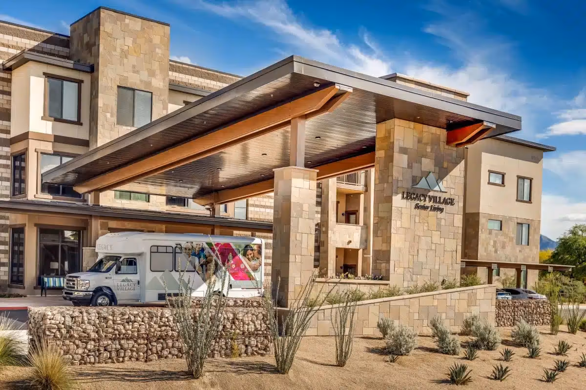 Exterior view of Legacy Village of Salt River senior living facility showing a modern building with stone and beige walls, a covered entrance with wooden beams, a shuttle bus parked under the entrance canopy, and desert landscaping with shrubs and rocks in the foreground under a blue sky with some clouds.