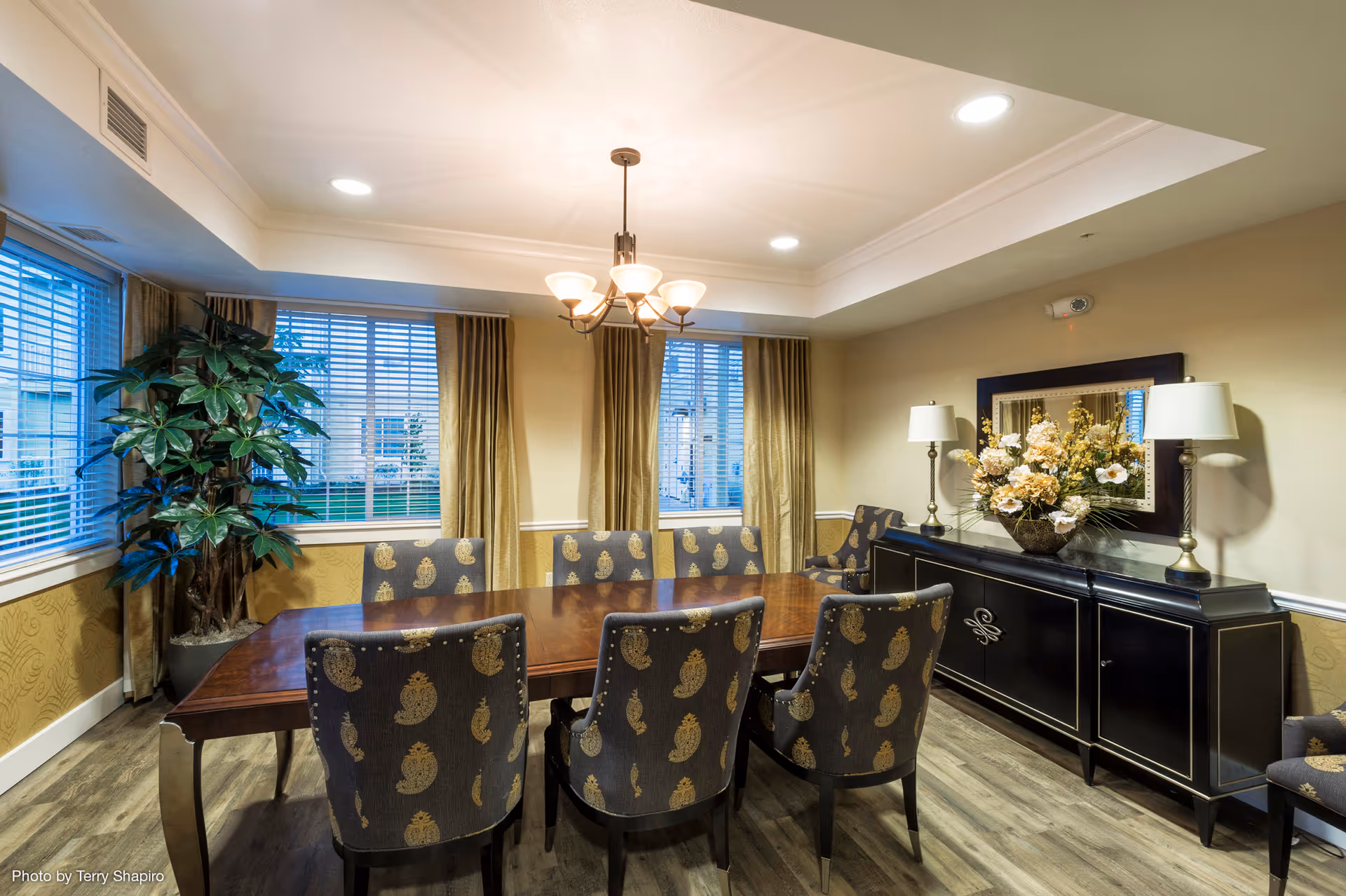 A well-lit dining room with a large wooden table surrounded by eight upholstered chairs featuring a paisley pattern. The room has three windows with blinds and gold curtains, a black sideboard with two lamps and a floral arrangement, and a chandelier hanging from the ceiling. There is a large green potted plant in the corner and wood flooring.