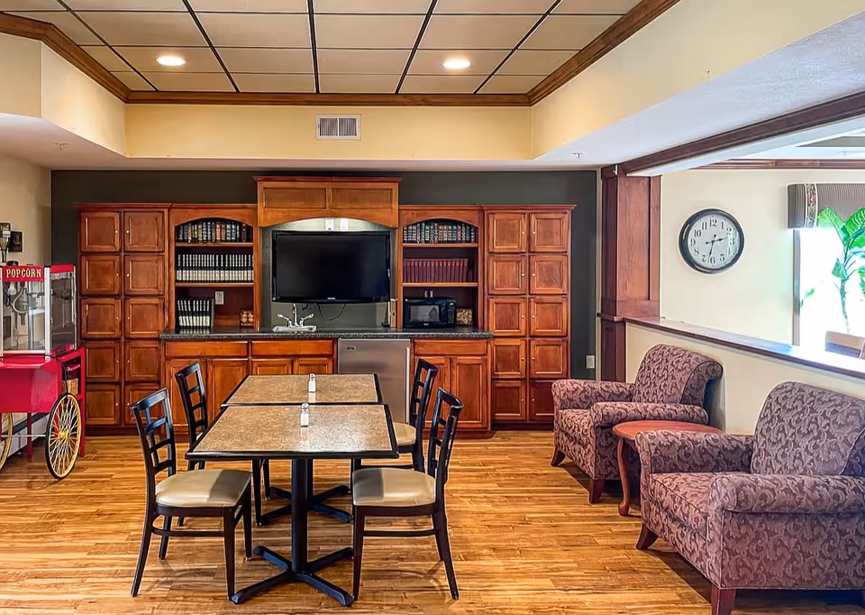 A cozy common area with a wooden floor and ceiling, featuring a table with four chairs in the center. Behind the table is a wooden cabinet with shelves holding books and a mounted flat-screen TV. To the right, there are two patterned armchairs with a small wooden side table between them. A popcorn machine is visible on the left side of the image, and a clock hangs on the wall to the right.