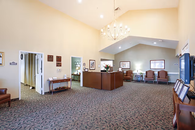 A spacious reception area with a high ceiling and chandelier. The room features a central wooden reception desk with flowers on top, several framed pictures on the walls, and a few armchairs arranged along the back wall. There are doorways leading to other rooms, and the floor is covered with patterned carpet.