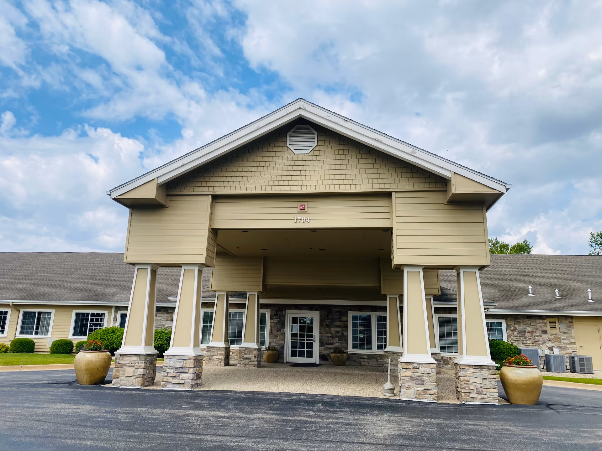 Front exterior view of a single-story building with a covered entrance supported by six columns. The building has beige siding, stone accents at the base of the columns, and multiple windows. Large planters with greenery are placed near the entrance. The sky is partly cloudy.