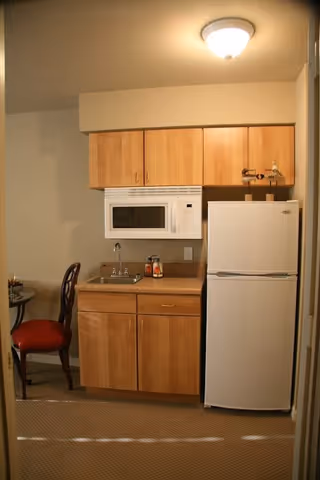 Small kitchenette area with wooden cabinets, a white microwave mounted above a countertop with a sink, and a white refrigerator. To the left, there is a partial view of a round table with a red cushioned chair.