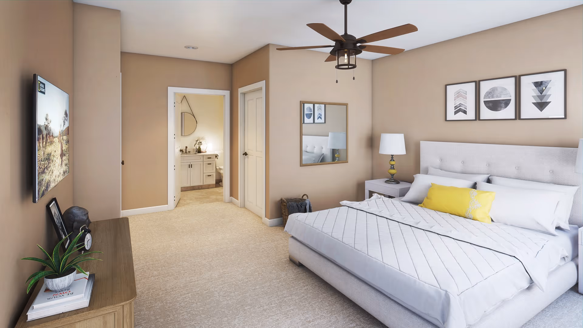 A bedroom with a large bed featuring white bedding and a yellow accent pillow. On the right wall above the bed are three framed geometric art pieces. A nightstand with a lamp is beside the bed. A ceiling fan with wooden blades hangs from the ceiling. On the left side, there is a dresser with a plant, a clock, and a framed photo. A flat-screen TV is mounted on the wall above the dresser. An open doorway leads to a bathroom with a vanity, mirror, and toilet visible. The walls are painted beige and the floor is carpeted.