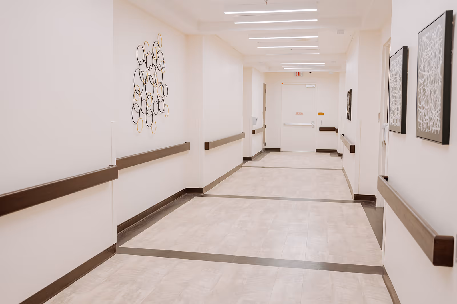 A clean, well-lit hallway in a healthcare facility with handrails on both sides, light-colored walls, and decorative wall art. The floor has a light tile pattern with darker borders, and there is an exit door at the end of the corridor.
