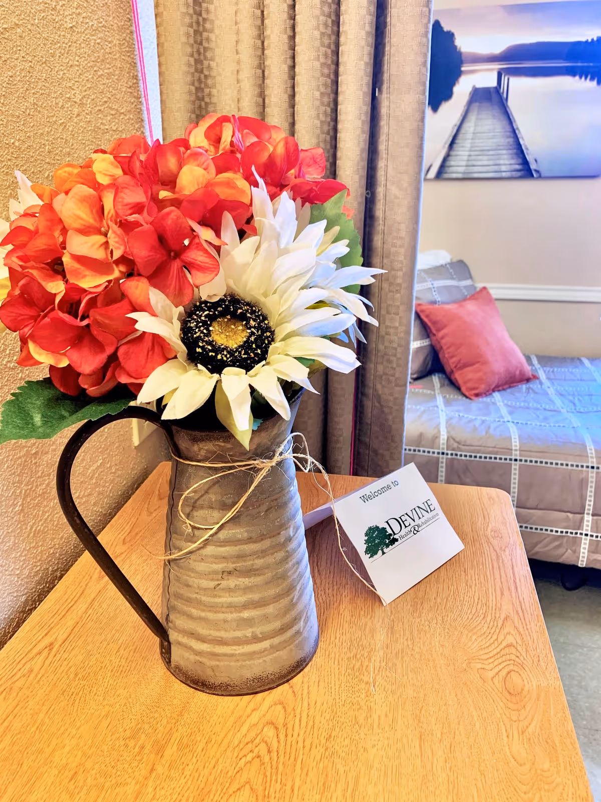 A close-up view of a rustic metal pitcher holding a bouquet of artificial flowers, including red and orange hydrangeas and a large white sunflower, placed on a wooden table. Behind the table, beige curtains hang next to a bed with a gray and white checkered bedspread and a red pillow. A framed picture of a dock extending into a lake is visible on the wall.