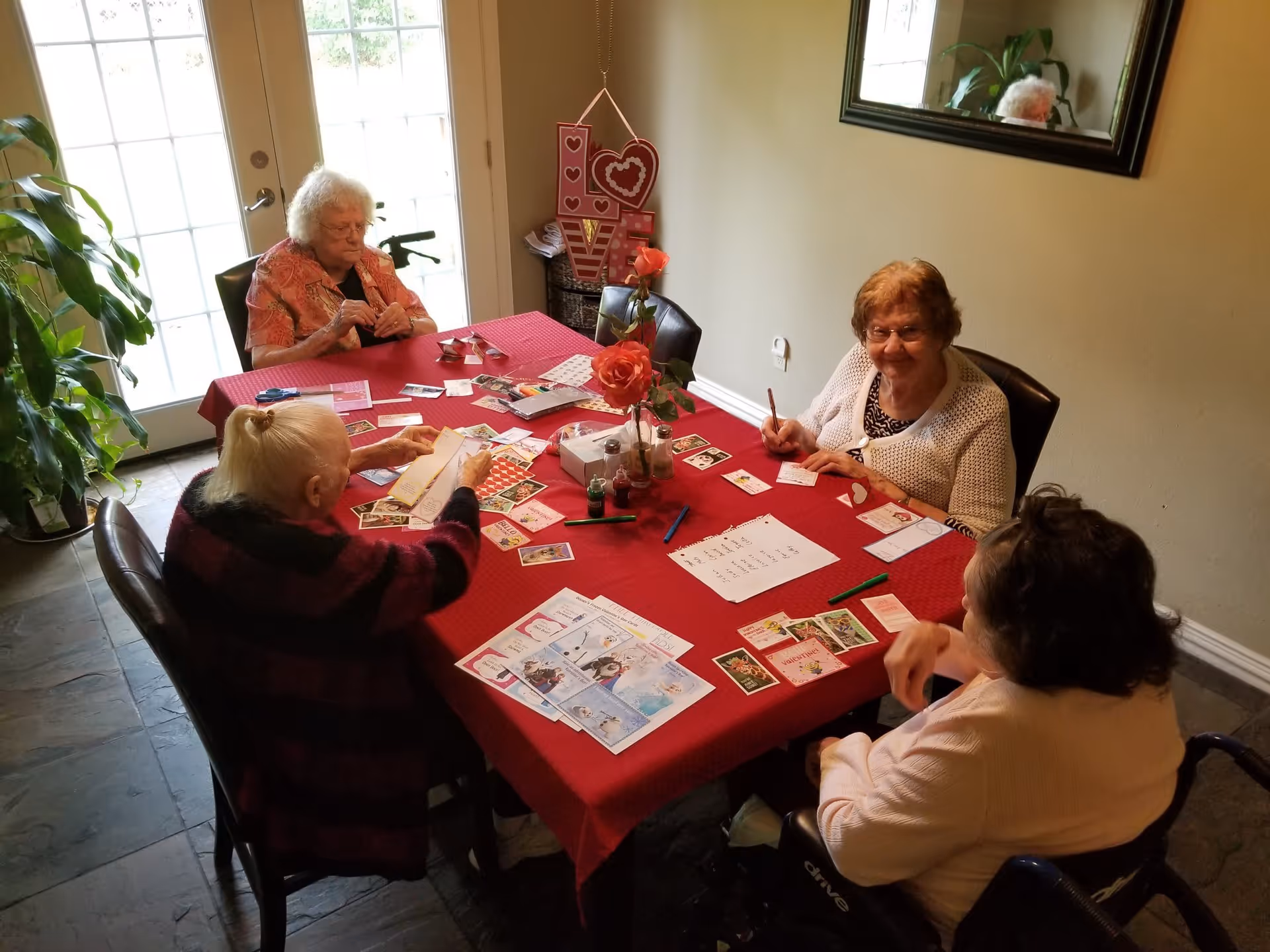 Four elderly women sitting around a rectangular table covered with a red tablecloth, engaging in a card or craft activity. The room has a large windowed door letting in natural light, a plant in the corner, a wall mirror, and a decorative 'LOVE' sign with hearts on the wall.