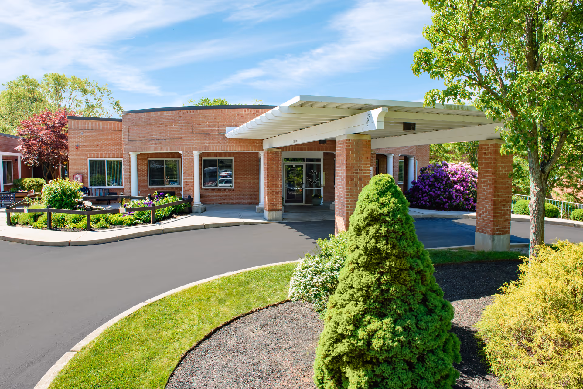 Exterior view of a brick building with a covered entrance and a circular driveway. The area is landscaped with green bushes, trees, and flowering plants under a blue sky with some clouds.