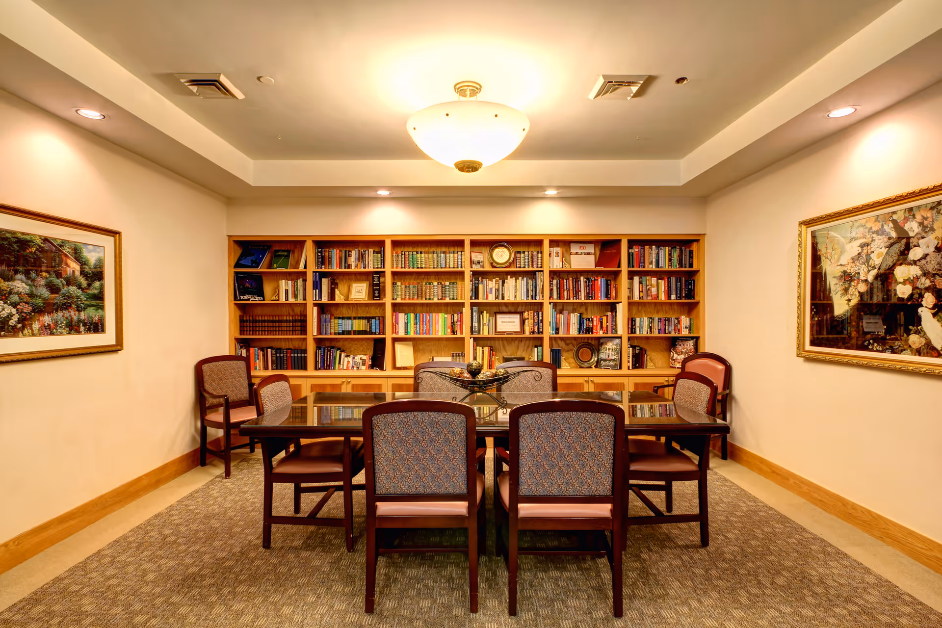 A cozy reading or meeting room with a large wooden bookshelf filled with books and decorative items against the back wall. In front of the bookshelf is a rectangular glass-top table surrounded by six cushioned chairs with wooden frames. The room is warmly lit by a ceiling light fixture and recessed lighting. Two framed paintings hang on the side walls.
