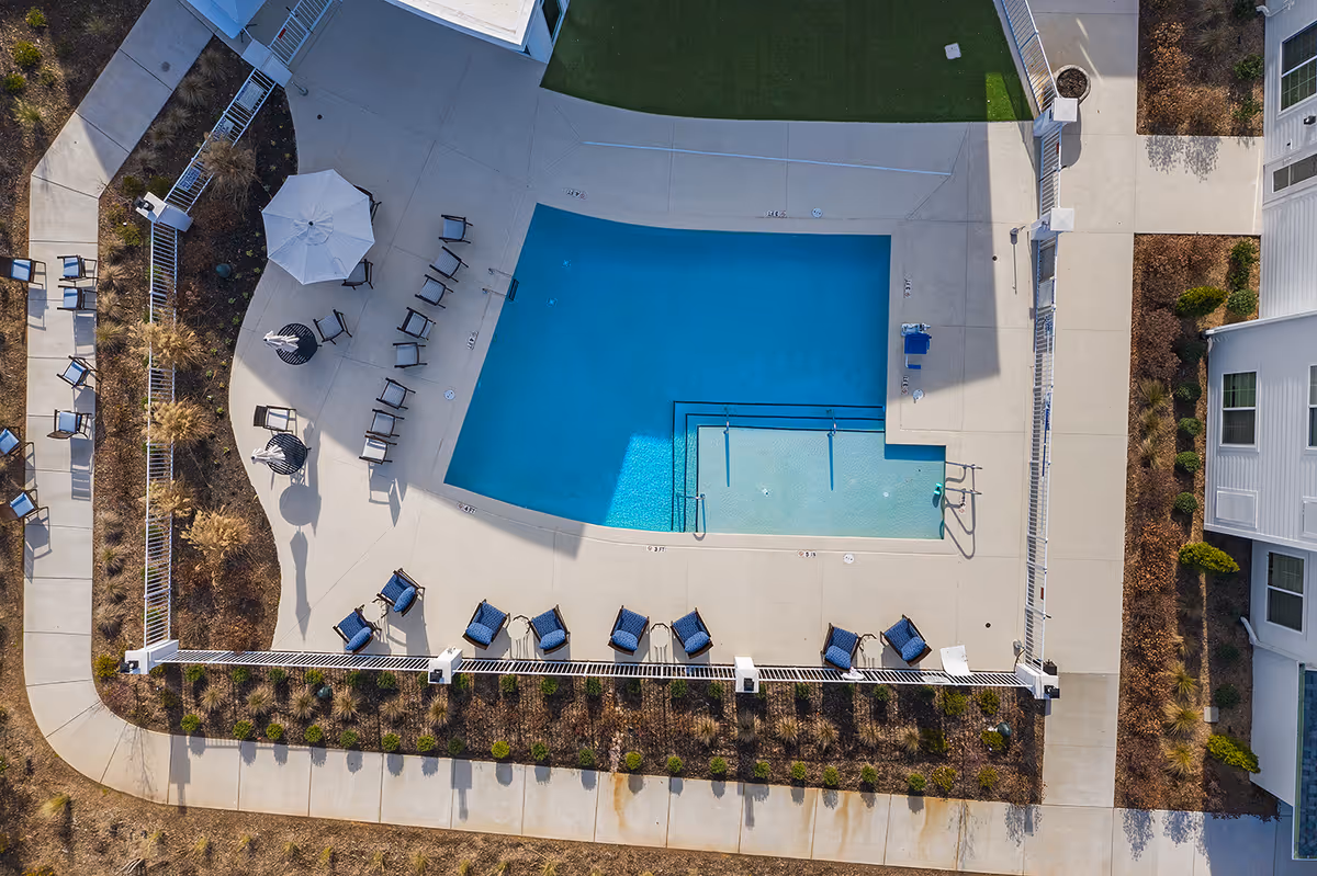 Aerial view of an outdoor swimming pool area at a senior living facility, featuring a rectangular pool with a shallow section, surrounded by lounge chairs, umbrellas, and a concrete deck. The pool area is enclosed by a white fence and adjacent to a building with windows.