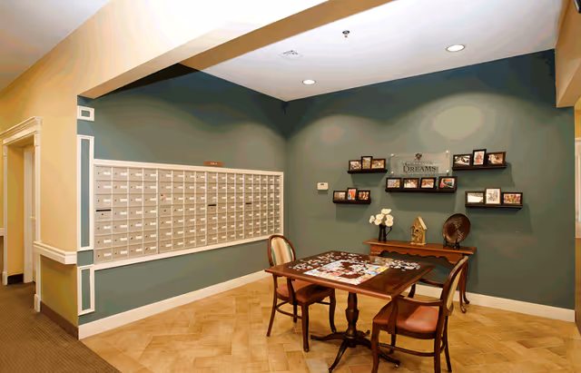 A cozy interior corner of a senior living facility featuring a wall of mailboxes on the left and a small wooden table with two chairs in the center. The table has a partially completed jigsaw puzzle on it. On the right wall, there are several small shelves displaying framed photos and decorative items, including a sign that reads 'Garden of Dreams'. The walls are painted in a muted green color with white trim, and the floor has a light wood pattern.