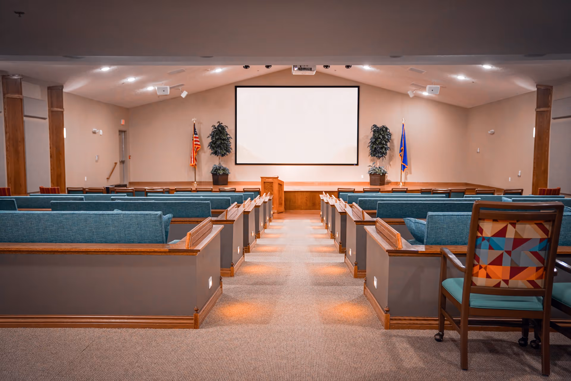Interior view of a meeting or presentation room with rows of blue cushioned benches facing a large blank projection screen. There are two potted plants on either side of the screen and two flags behind the stage area. The room has carpeted flooring and a high ceiling with recessed lighting.