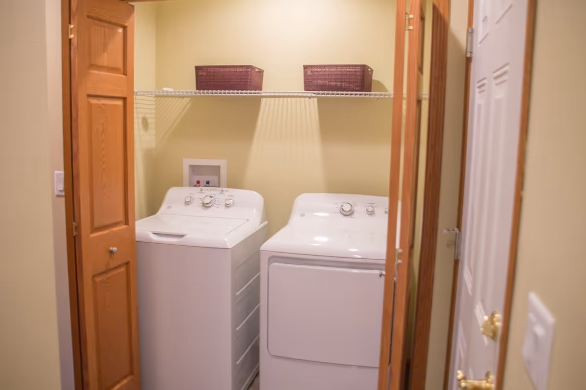 Small laundry room with a white washing machine and dryer side by side, a wire shelf above holding two purple baskets, and wooden bi-fold doors partially open.