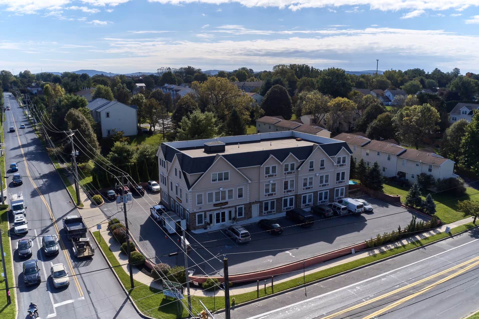 Aerial view of a three-story residential building named Alexandria Manor surrounded by trees and houses, with a parking lot in front and roads on two sides.