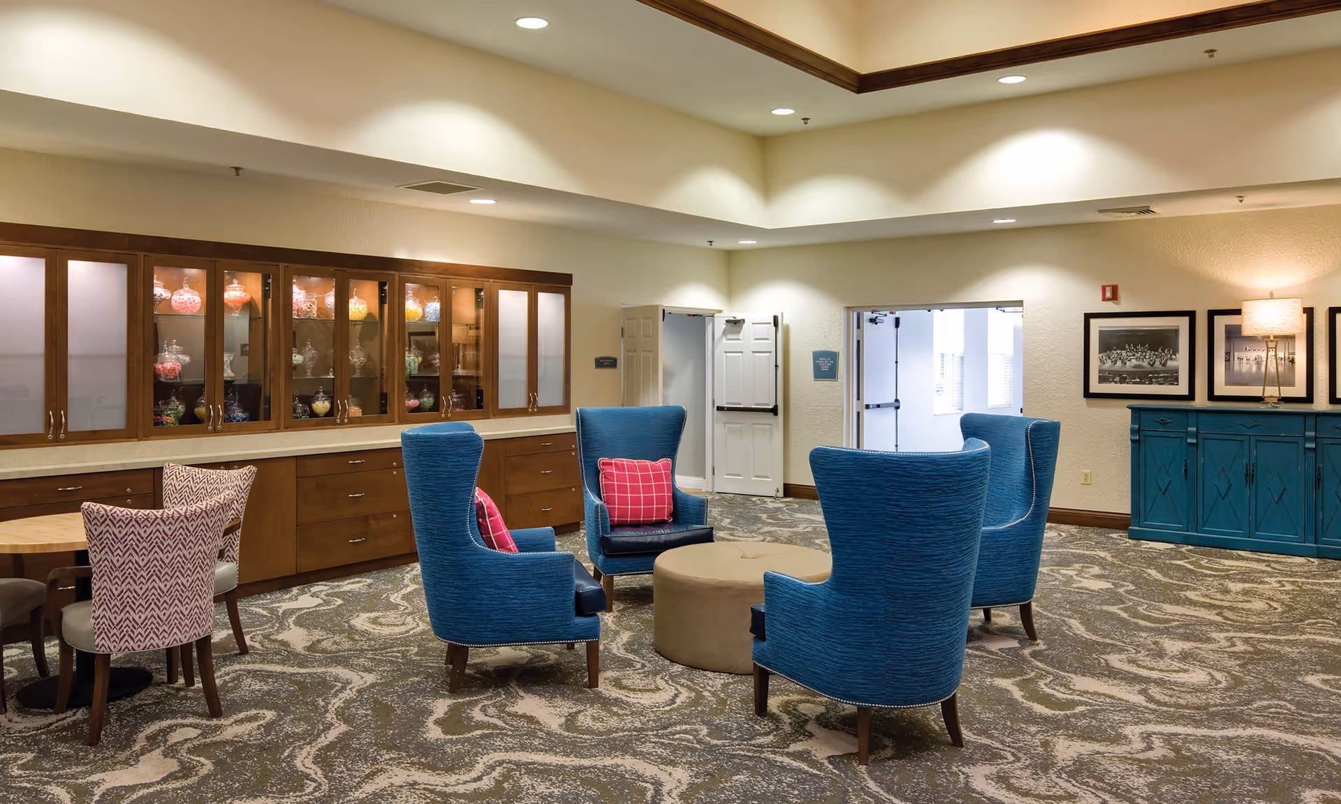 A cozy common area in a senior living facility featuring four blue upholstered armchairs with pink pillows arranged around a round beige ottoman. To the left, there is a wooden cabinet with glass doors displaying jars filled with colorful candies. A small round table with patterned chairs is also visible. The room has patterned carpet flooring, framed black and white photos on the wall, and soft overhead lighting.