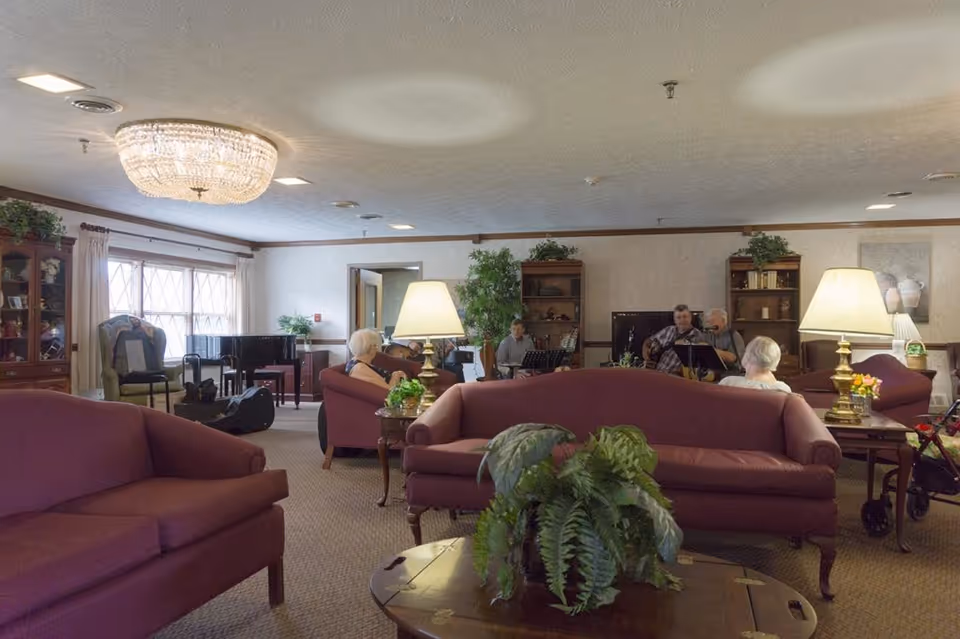 A cozy living room in a senior living facility with maroon sofas, a coffee table with a green plant, two table lamps, and a group of elderly people seated and listening to musicians playing guitar and keyboard in the background. The room has large windows with curtains, wooden bookshelves, and a chandelier on the ceiling.