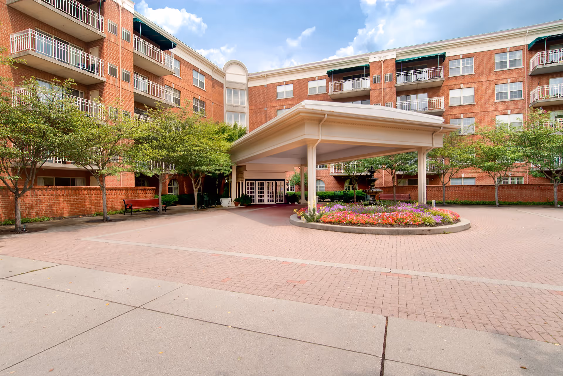 Entrance area of a senior living facility with a covered drop-off zone surrounded by a circular flower bed with colorful flowers. The building is made of red brick with multiple balconies and windows. There are small trees and benches along the sides of the entrance area under a partly cloudy sky.