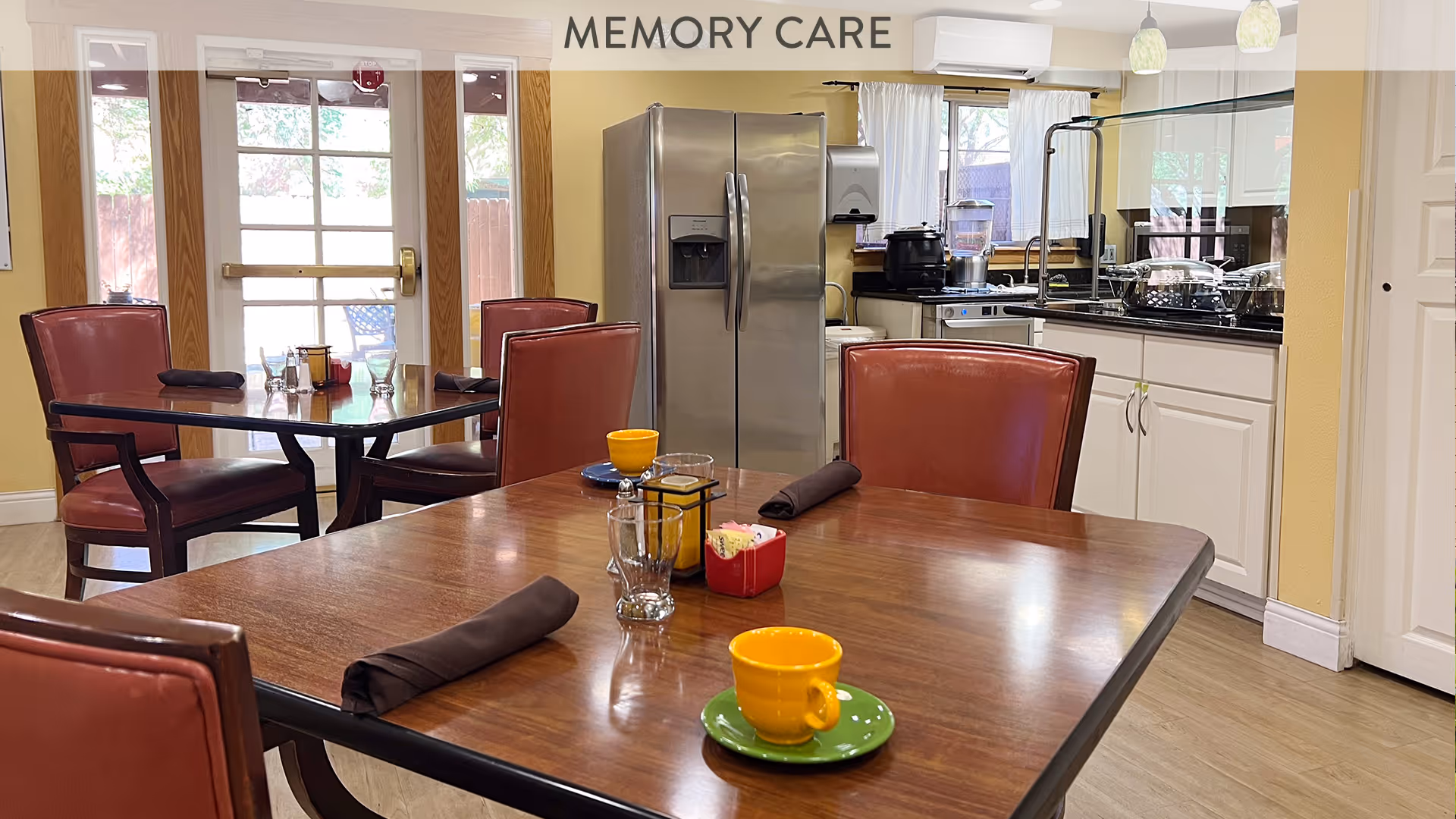 A cozy dining area in a memory care facility with wooden tables and red cushioned chairs. The tables are set with yellow cups on green saucers, glasses, napkins, and condiments. In the background, there is a stainless steel refrigerator, kitchen counters with appliances, and large windows letting in natural light.