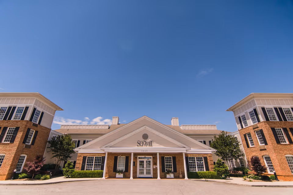 Front exterior view of The Summit senior living facility building with a clear blue sky above. The building features a symmetrical design with brick and white siding, multiple windows, and a central entrance with a covered porch supported by white columns.