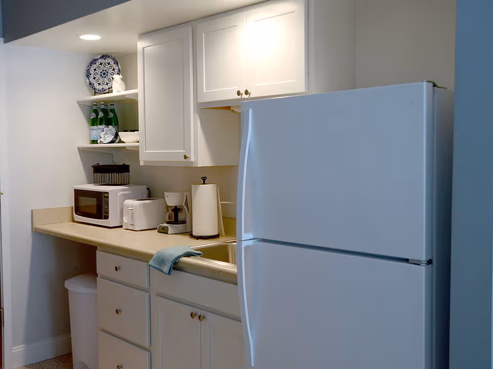 A small kitchen area with white cabinets and beige countertops. The kitchen includes a white refrigerator, a microwave, a toaster, a coffee maker, a paper towel holder, and a sink with a blue towel draped over the edge. There are decorative plates and bottles on open shelves above the counter.
