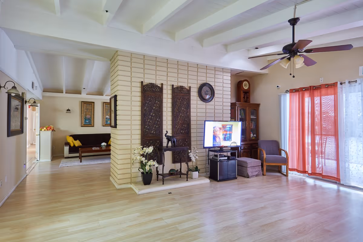 A spacious living room area with light wood flooring and a white beamed ceiling. The room features a brick partition wall decorated with wooden panels and plants. There is a TV on a stand, a wooden cabinet with a clock on top, a gray armchair with an ottoman, and red curtains covering a sliding glass door. In the background, another seating area with a dark sofa and yellow pillows is visible.