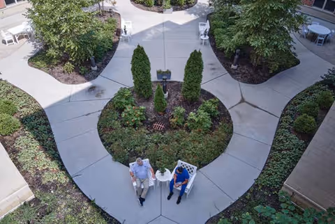 Overhead view of an outdoor courtyard area with concrete walkways surrounding landscaped garden beds. Two people are seated on white chairs at a small white table in the center of the courtyard. There are additional white chairs and tables around the perimeter near the building walls.