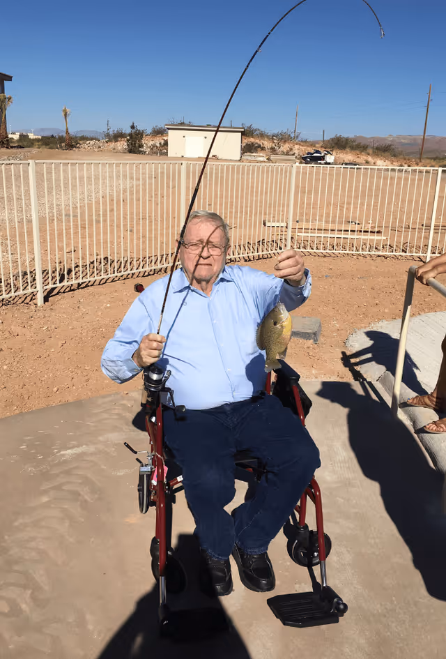 An elderly man sitting in a wheelchair outdoors holding a fishing rod with a fish he caught. He is wearing a light blue shirt and dark pants. The background shows a fenced area with a dirt ground and a small building, under a clear blue sky.