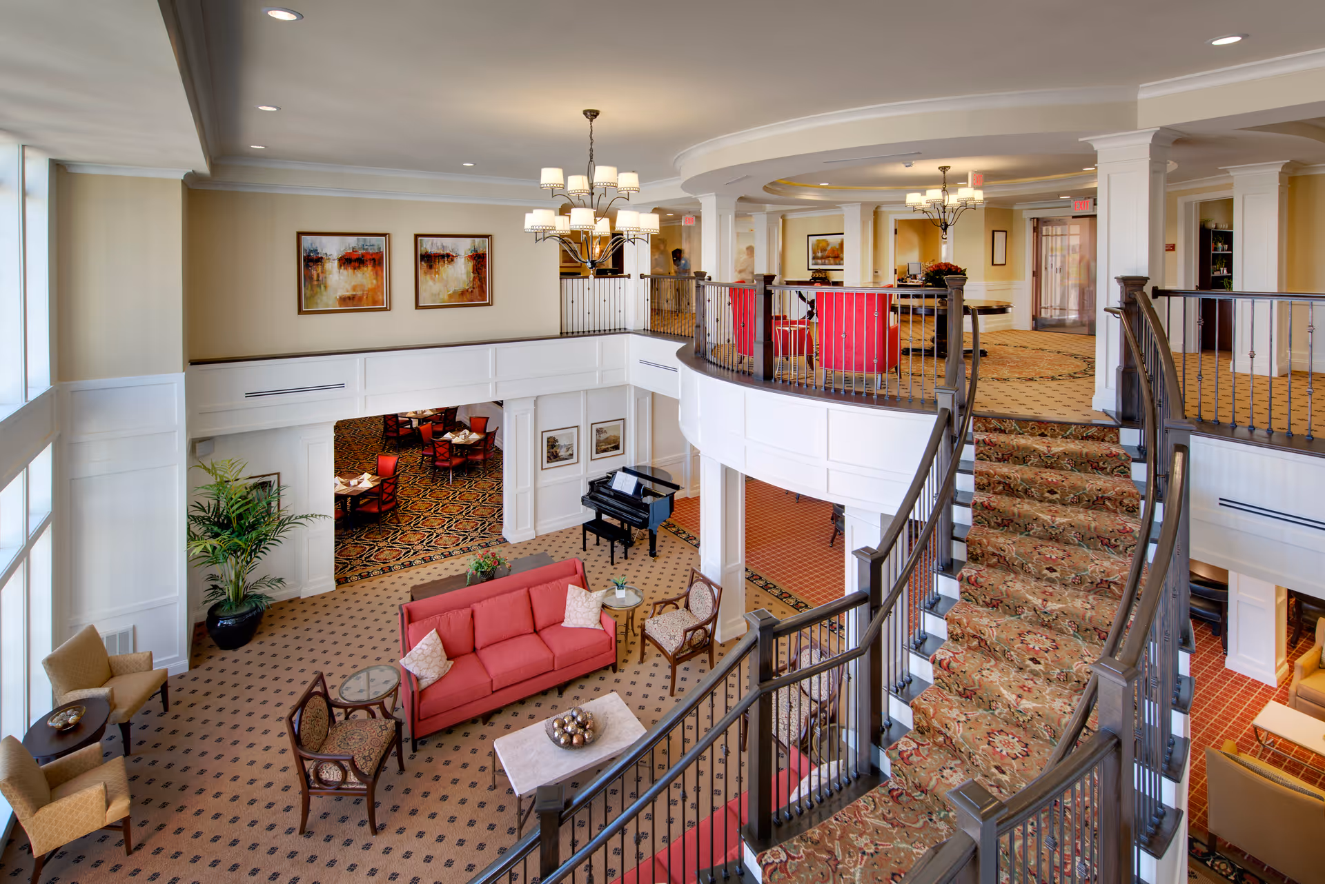 Interior view of a senior living facility featuring a spacious two-level common area with a carpeted staircase, red upholstered chairs and sofa, a black grand piano, and decorative paintings on the walls. The upper level has a seating area with red chairs and chandeliers hanging from the ceiling.