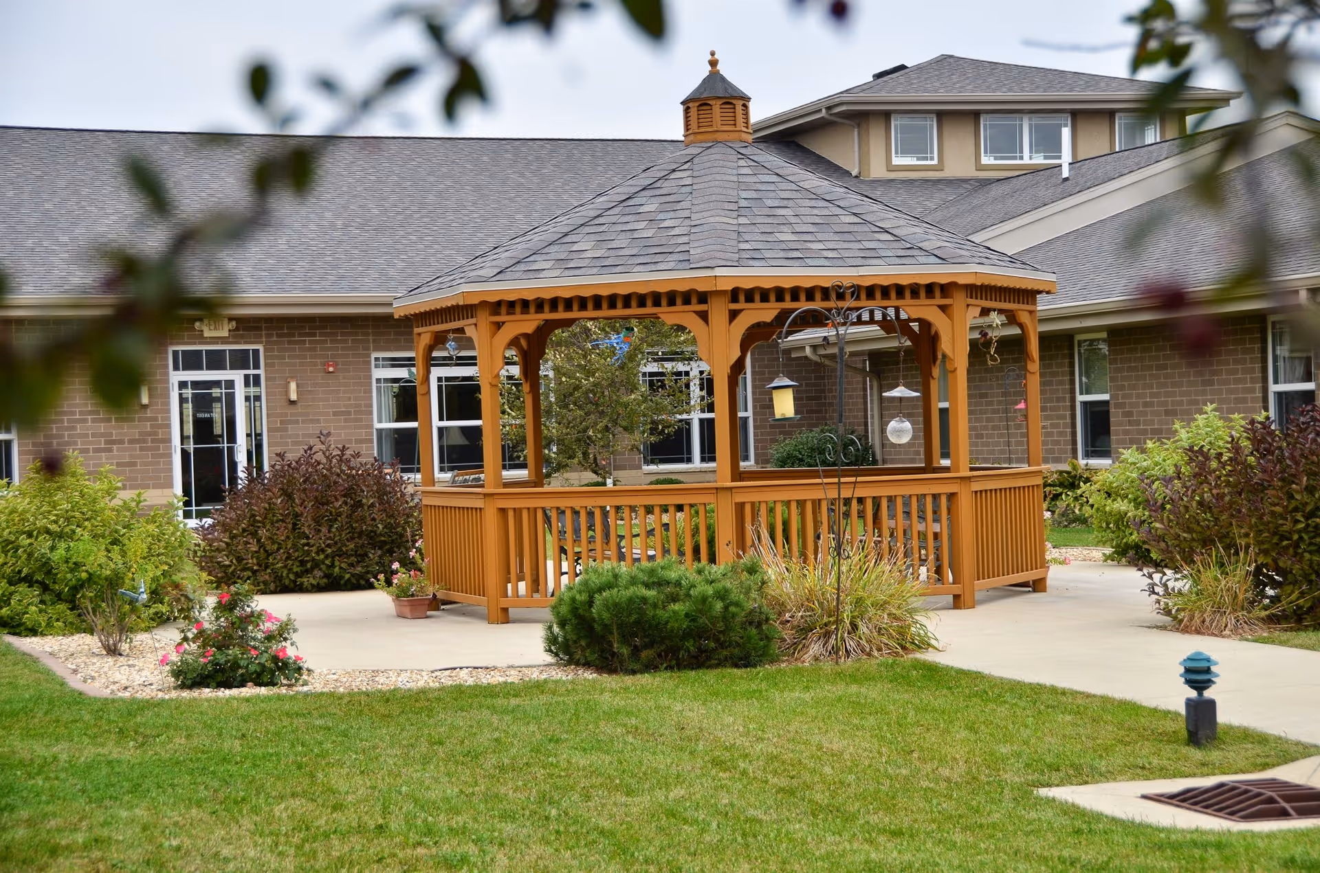 A wooden gazebo with a shingled roof situated in a landscaped garden area with green grass, shrubs, and flowers. The gazebo is surrounded by a concrete walkway and is in front of a brick building with multiple windows and doors.