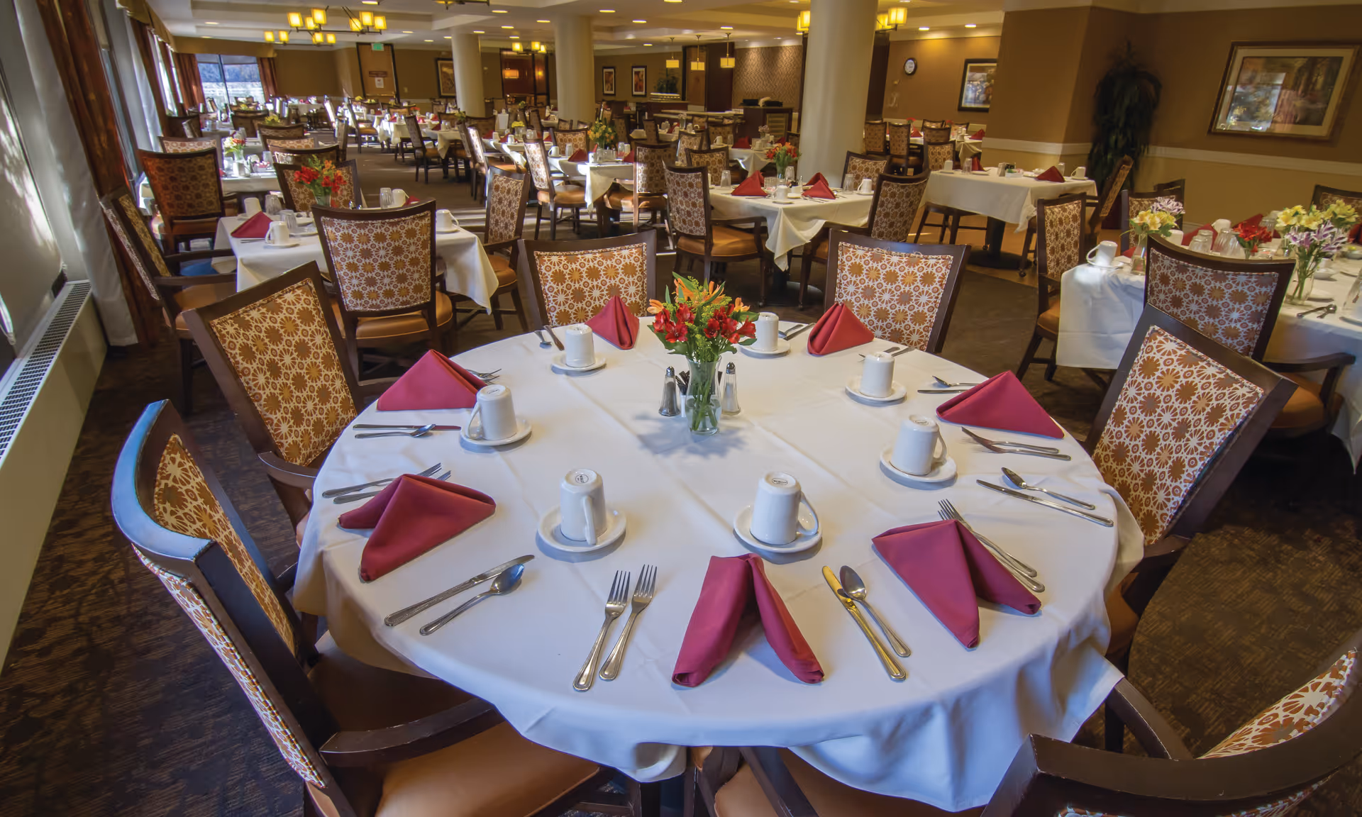 A spacious dining room with round tables set with white tablecloths, red folded napkins, cups, silverware, and patterned chairs.
