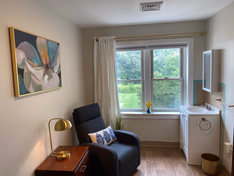 Cozy room with a reclining chair by a window, a side table with a lamp, wall art, and a small sink/vanity.