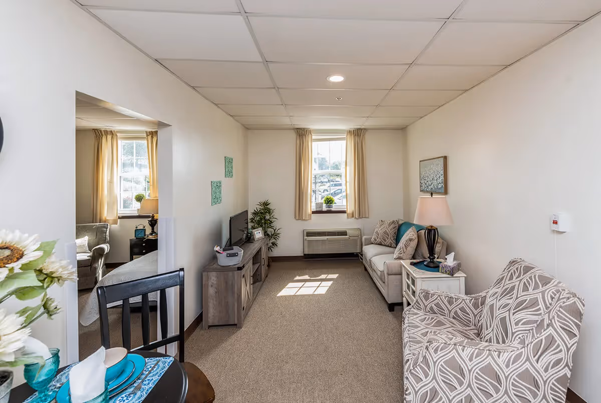 A cozy living room in a senior living facility with beige walls and carpet. The room features a patterned armchair, a beige sofa with decorative pillows, a side table with a lamp and tissues, a TV on a wooden stand, and a window with beige curtains letting in natural light. A small dining table with place settings and flowers is partially visible in the foreground, and a bedroom with a bed and armchair is visible through an open doorway.