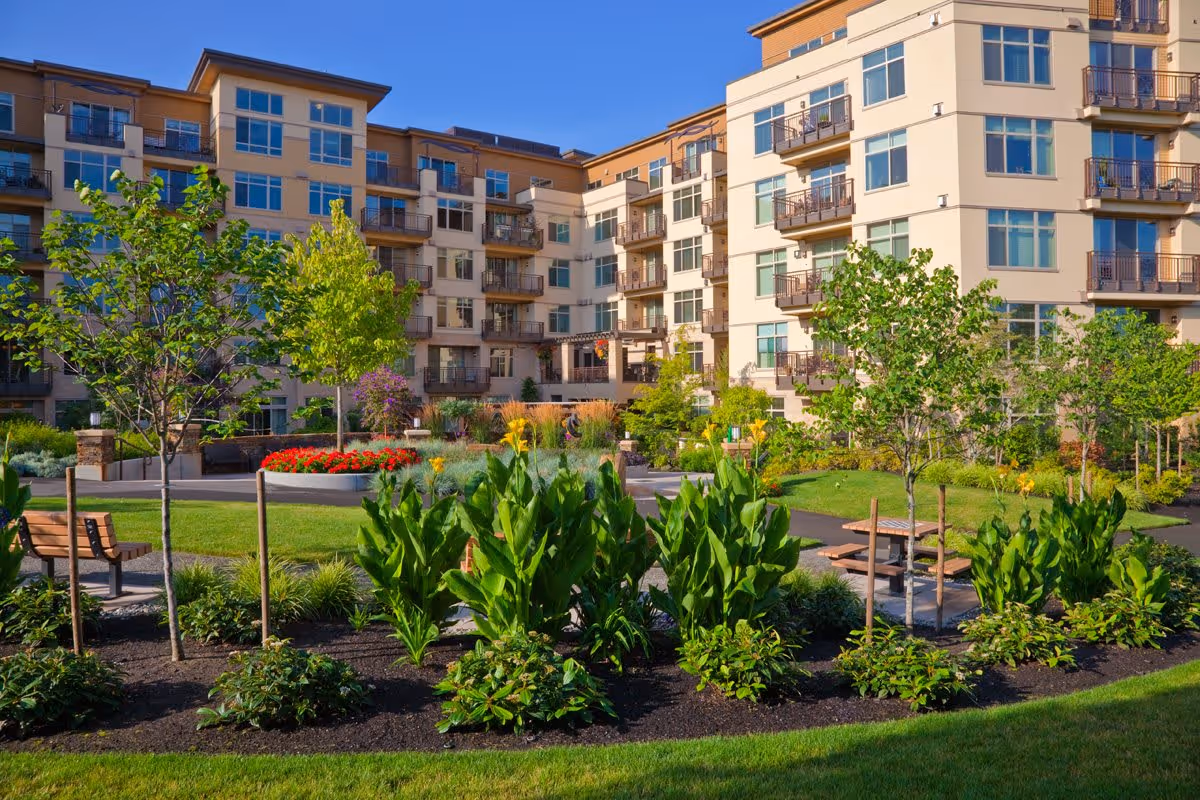 A landscaped outdoor garden area with green plants, trees, and flowers in front of a multi-story residential building with balconies under a clear blue sky.