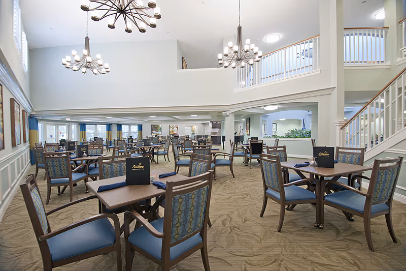 Spacious dining room in a senior living facility with multiple wooden tables and chairs arranged neatly. The room features high ceilings with modern chandeliers, large windows with blue and yellow curtains, and a staircase leading to an upper level. Menus labeled Allegro are placed on some tables.