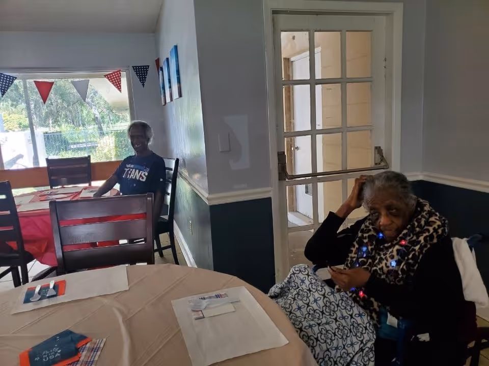Two elderly people sit at decorated dining tables in a communal room with patriotic bunting and a glass-paned door.